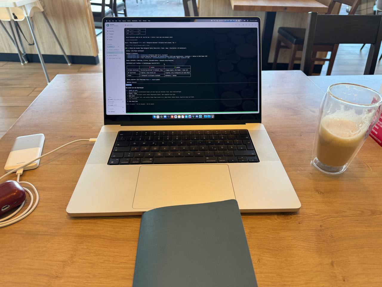 MacBook in einem Café mit Kaffee daneben. Auf dem Bildschirm ist ein dunkles Terminalfenster zu sehen; Remo arbeitet während einer Wartezeit an Packliste.